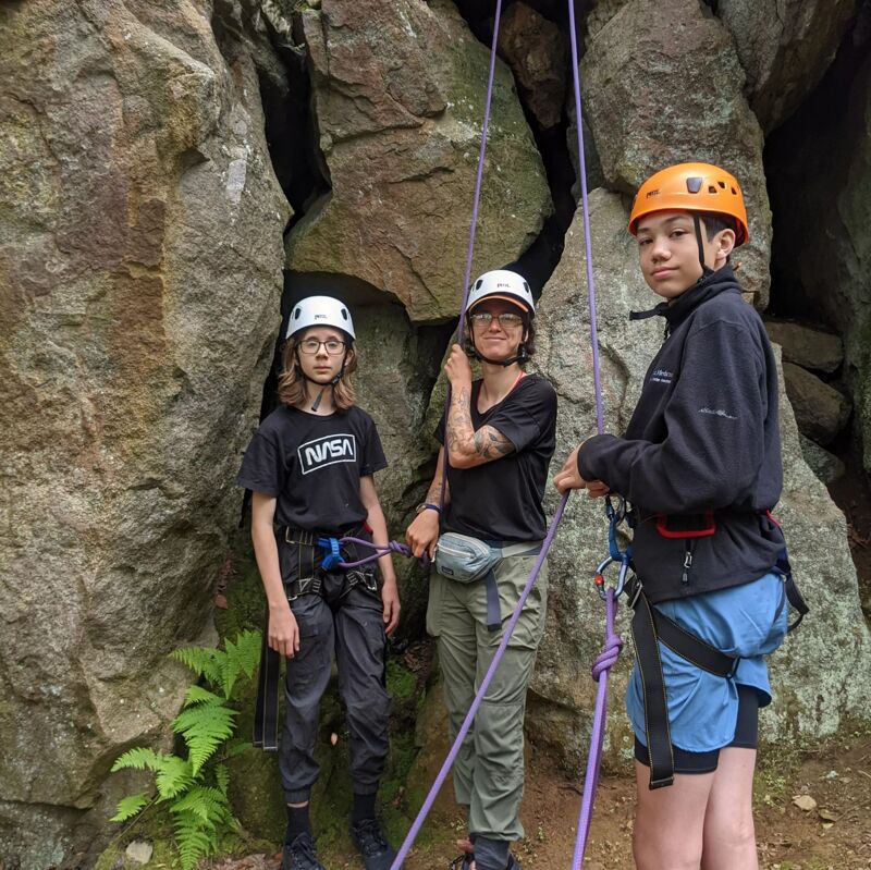 Three young individuals are geared up for rock climbing, standing in front of a rocky cliff face. They are all wearing helmets and harnesses, with climbing ropes visible. The person on the left wears a NASA t-shirt, while the person in the middle has tattoos on their arm. The person on the right is wearing a black jacket and blue shorts. Ferns grow at the base of the rocks, suggesting a natural climbing environment.